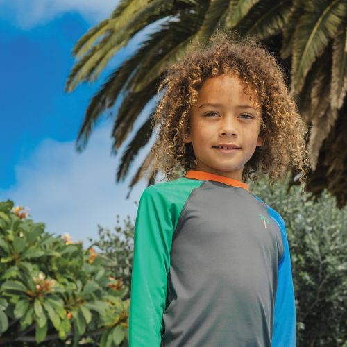 A young child with curly hair wearing a long-sleeve athletic shirt with green, blue, and gray panels and an orange collar, photographed outdoors against palm leaves and blue sky.