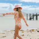 A young girl wearing a pink sun hat and a long-sleeve floral swimsuit walks along a sandy beach with the ocean and a wooden pier in the background.