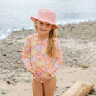 A young girl stands on a sandy, rocky beach wearing a pink floral long-sleeve swim shirt and a matching pink bucket hat.