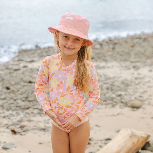 A young girl stands on a sandy, rocky beach wearing a pink floral long-sleeve swim shirt and a matching pink bucket hat.