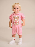 A toddler with blonde hair stands against a solid beige studio background wearing a pink and white striped short-sleeve romper with a teddy bear graphic on the front, paired with white socks and white sneakers.