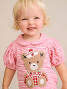 A smiling toddler with blonde hair wearing a pink short-sleeve striped shirt with a teddy bear applique on the front, photographed against a plain beige background.