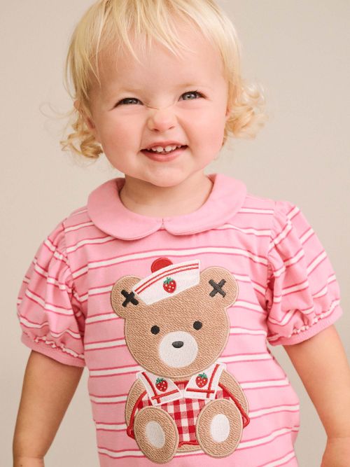 A smiling toddler with blonde hair wearing a pink short-sleeve striped shirt with a teddy bear applique on the front, photographed against a plain beige background.