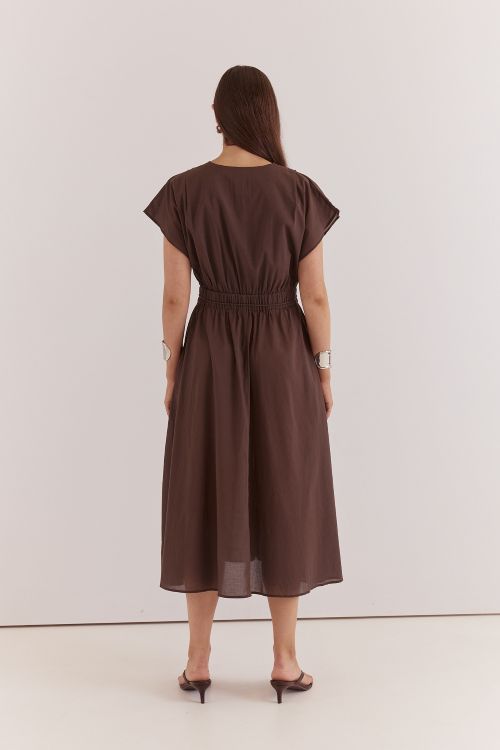 A woman viewed from the back wearing a dark brown midi dress with short flutter sleeves, a round neckline, and a gathered elastic waist, paired with dark heeled sandals against a plain light gray studio background.