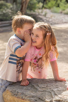 Two children sitting outdoors on a stone surface, with the girl wearing a pink short-sleeve shirt with a cowboy and horse graphic and the boy wearing a white graphic T-shirt. The background shows a blurred outdoor setting with trees and sunlight.