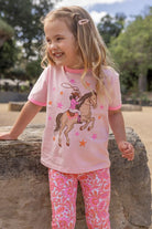 A young girl with blonde hair is smiling outdoors, wearing a light pink short-sleeve t-shirt with a cowboy on a horse graphic and pink patterned leggings.