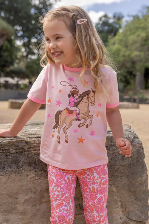 A young girl with blonde hair is smiling outdoors, wearing a light pink short-sleeve t-shirt with a cowboy on a horse graphic and pink patterned leggings.
