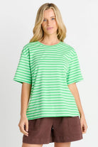 A woman modeling a green and white horizontally striped short-sleeve t-shirt with brown shorts, set against a plain light gray studio background.