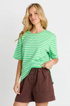 A woman modeling a green and white striped short-sleeve t-shirt paired with brown shorts, set against a plain light gray studio background.