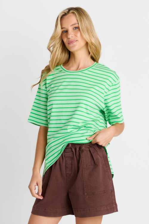 A woman modeling a green and white striped short-sleeve t-shirt paired with brown shorts, set against a plain light gray studio background.