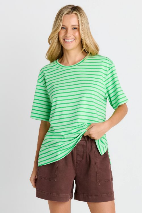 A woman modeling a bright green short-sleeve t-shirt with thin white horizontal stripes, paired with brown shorts, against a light gray studio background.