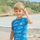 A young boy with blonde hair stands on a beach, holding a seashell to his ear. He is wearing a bright blue short-sleeve swim shirt with a white fish pattern.
