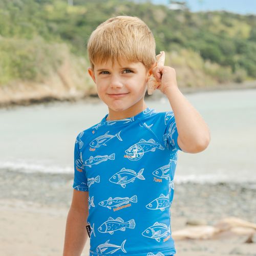 A young boy with blonde hair stands on a beach, holding a seashell to his ear. He is wearing a bright blue short-sleeve swim shirt with a white fish pattern.