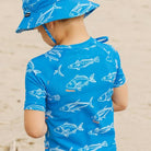 A child wearing a bright blue rash guard and matching sun hat with a white fish pattern, photographed from behind on a sandy beach.