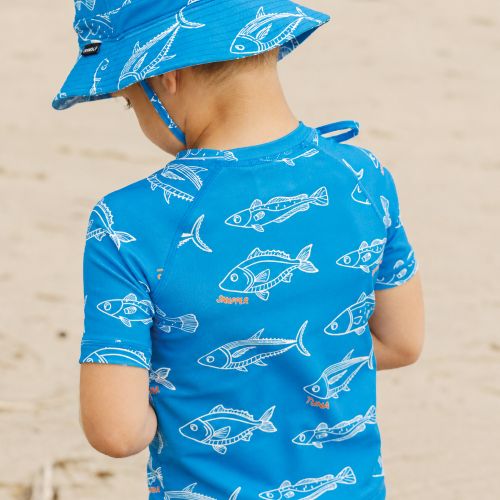 A child wearing a bright blue rash guard and matching sun hat with a white fish pattern, photographed from behind on a sandy beach.