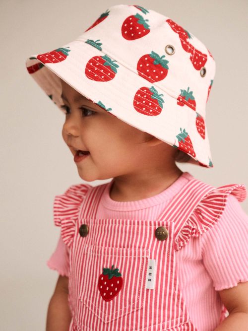 A toddler wearing a white bucket hat with a red strawberry print and a pink striped ruffle-shoulder top, shown in profile against a plain light background.