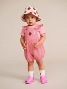 A toddler standing against a plain beige studio background, wearing a pink strawberry-print sun hat, pink and white striped overall romper with ruffled sleeves, white socks, and pink clogs.