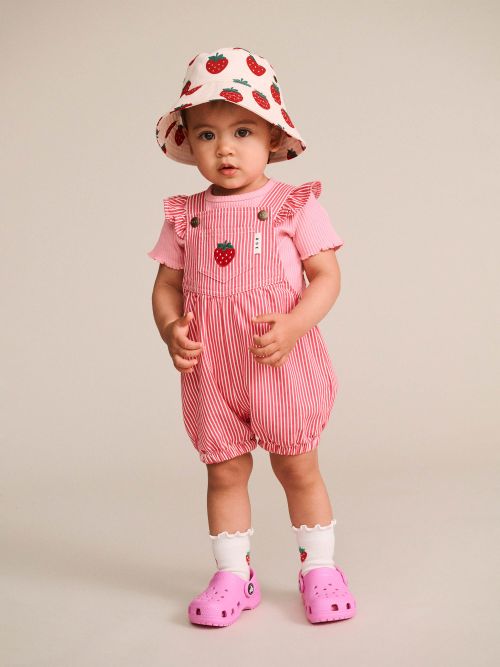 A toddler standing against a plain beige studio background, wearing a pink strawberry-print sun hat, pink and white striped overall romper with ruffled sleeves, white socks, and pink clogs.