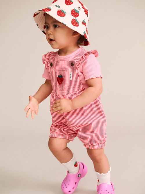 A toddler wearing a pink and white striped short-sleeve romper with ruffled shoulders, a matching strawberry-print sun hat, white socks, and pink clogs, posed against a plain light beige studio background.