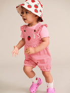 A toddler wearing a pink and white striped short-sleeve romper with ruffled shoulders, a matching strawberry-print sun hat, white socks, and pink clogs, posed against a plain light beige studio background.