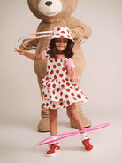 A young girl wearing a strawberry-print dress, matching hat, and red sneakers poses in front of a giant teddy bear while spinning a pink hula hoop on a light beige studio background.