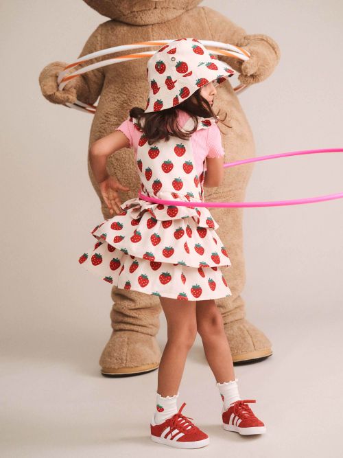 A young girl wearing a strawberry-print tiered dress, matching hat, and red sneakers stands in front of a large teddy bear costume holding hula hoops, photographed against a plain light beige studio background.