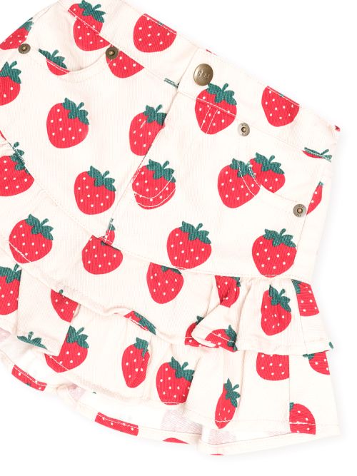 A close-up of a ruffled skirt with a red strawberry print on a cream background, featuring button details and pockets.