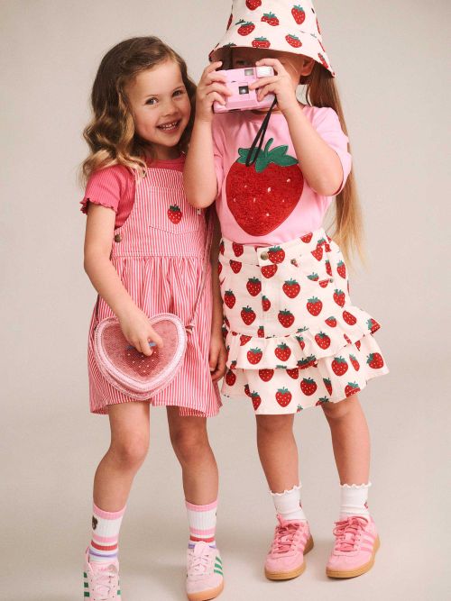 Two young girls modeling strawberry-themed clothing against a plain beige studio background. The girl on the left wears a pink and white striped pinafore dress with a strawberry motif, holding a pink heart-shaped accessory.