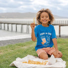 A smiling child with curly hair sits on grass by a waterfront pier, holding up a french fry. The child wears a bright blue graphic T-shirt that reads 'FISH 'N CHILI SIMPLY MY FAVORITE' with an orange-and-white checkered shorts, with a paper tray of fries in front.