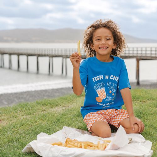 A smiling child with curly hair sits on grass by a waterfront pier, holding up a french fry. The child wears a bright blue graphic T-shirt that reads 'FISH 'N CHILI SIMPLY MY FAVORITE' with an orange-and-white checkered shorts, with a paper tray of fries in front.