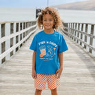 A smiling child standing on a wooden boardwalk by the water, wearing a bright blue graphic T-shirt that reads “FISH ’N CHIPS” with a fish and fries illustration, paired with orange-and-white checkered shorts.