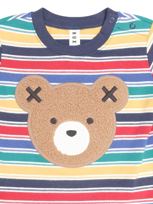 A close-up of a children's striped long-sleeve top with a large brown teddy bear face applique on the front. The shirt features horizontal stripes in navy blue, red, yellow, green, and white, with a navy collar and a white tag reading 'XUX' inside the neck.