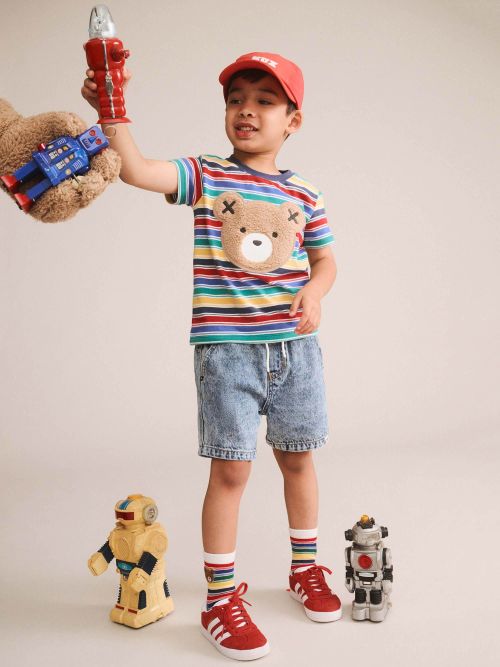 A young boy modeling a colorful striped t-shirt with a teddy bear face graphic, paired with denim shorts, red sneakers, and a red cap. He is holding a toy robot in his raised hand, with small toy robots on the floor against a plain beige studio background.