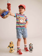 A young boy modeling a colorful striped t-shirt with a teddy bear face graphic, paired with denim shorts, red sneakers, and a red cap. He is holding a toy robot in his raised hand, with small toy robots on the floor against a plain beige studio background.
