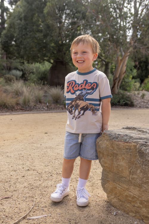 A young boy smiling outdoors next to a large rock, wearing a beige graphic t-shirt with 'Rodeo' and a cowboy print, blue denim shorts, white socks, and white sneakers.